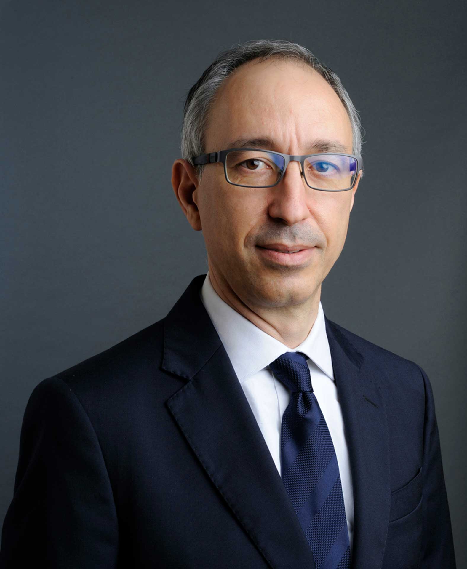 Man with short grey hair and glasses wearing a dark suit with a white shirt and tie